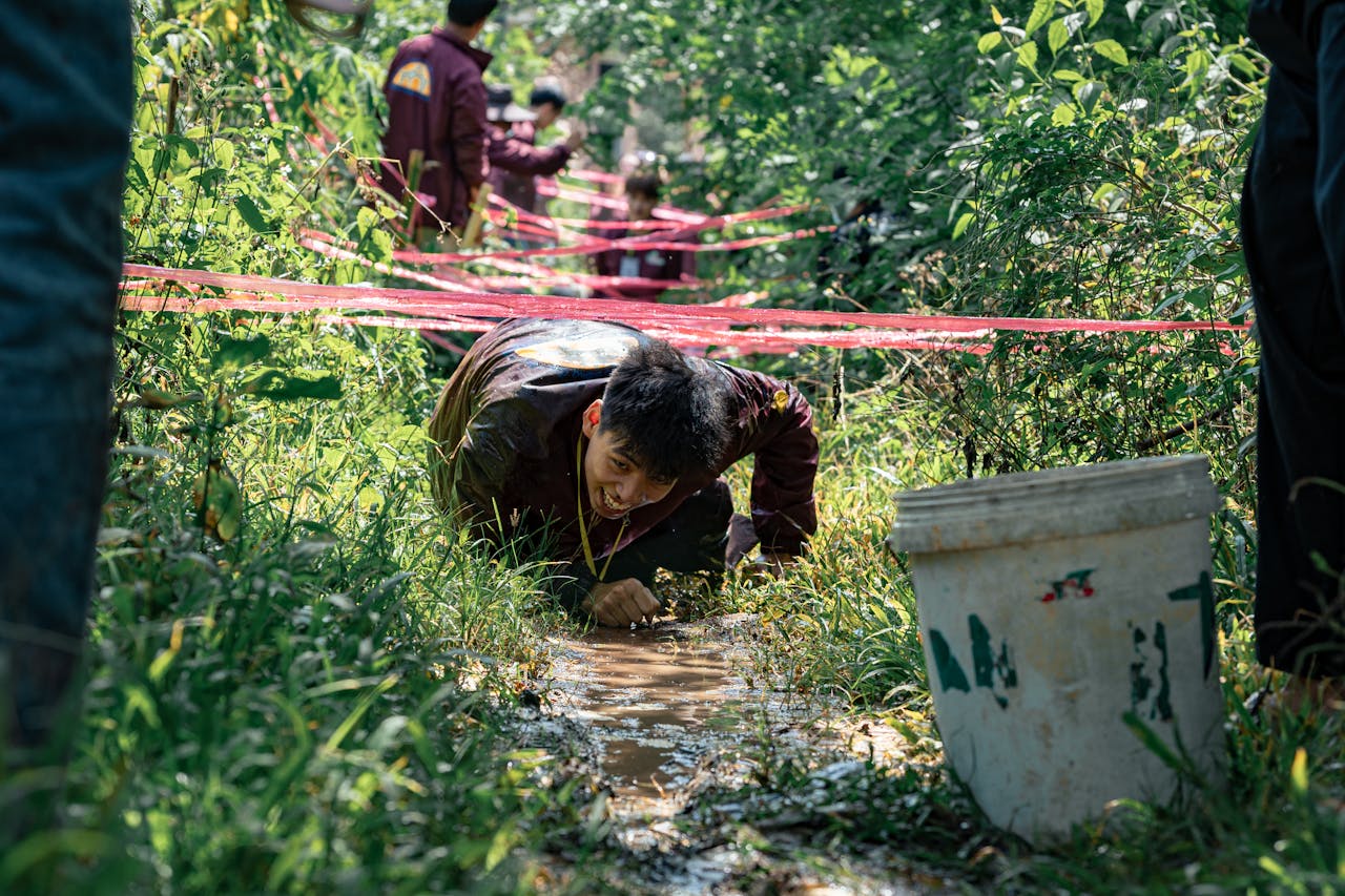 Teenager navigating a muddy obstacle course in a lush outdoor setting during a sunny day.